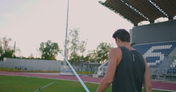 Oszczep Thrower spaceruje po stadionie z włóczniami i je zbiera. Idź z włócznią w ręku na tle stadionu stoi. Szkolenie mistrza olimpijskiego. — Wideo stockowe