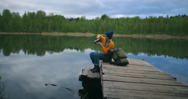 Kobieta w kapeluszu i plecaku siedzi na drewnianym pomoście leśnego jeziora w górach i pije gorące napoje herbata lub kawa z termosu. — Wideo stockowe