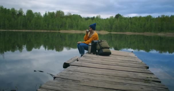 A female traveler drinks tea with a backpack and enjoys the beauty of a mountain forest lake sitting on a wooden pier. Slow-motion concept of a lonely womans solitary journey. Quarantine and travel — Stock Video