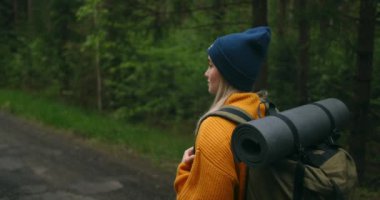 Arkadan ve aşağıdan bakıldığında, sarı bir kazak ve şapkayla orman yolunda sırt çantalı ve ağaçlara ve doğaya bakan bir kadın görülüyor. Yolculukta yalnızdım. Özgürlük kavramı 