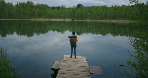 Back view: A female traveler with a backpack enjoys the beauty of a mountain forest lake standing on a wooden pier. Slow-motion concept of a lonely womans solitary journey. Quarantine and travel — Stock Video