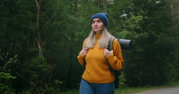 黄色のセーターと帽子のバックパックを持つ女性の肖像画低角度スローモーションで森の中の道路に沿って歩いている. — ストック動画