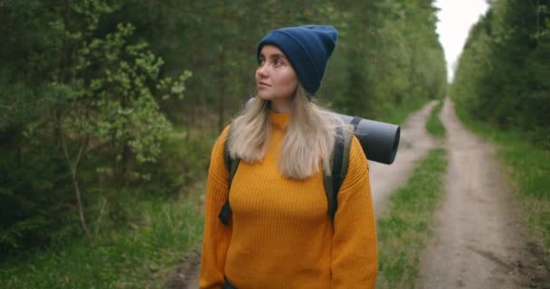 Uma mulher viaja a pé em uma camisola e chapéu ao longo de uma estrada florestal com uma mochila e examina as árvores e a natureza. Sozinho numa viagem. O conceito de liberdade e viagens . — Vídeo de Stock