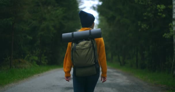 Pandangan kembali dari bawah dan samping: seorang wanita berjalan kaki dengan sweater kuning dan topi di jalan hutan dengan ransel dan melihat pohon dan alam. Sendirian di perjalanan. Konsep kebebasan. — Stok Video