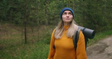 Alçak açılı bir portre. Sırt çantalı, sarı kazaklı ve şapkalı bir kadın ormanda ağır çekimde yol boyunca yürüyor..