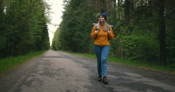 Vista frontal, plano principal: uma mulher viajando a pé em uma camisola amarela e chapéu em uma estrada florestal com uma mochila e vê as árvores e a natureza. Sozinho numa viagem. O conceito de liberdade e viagens . — Vídeo de Stock