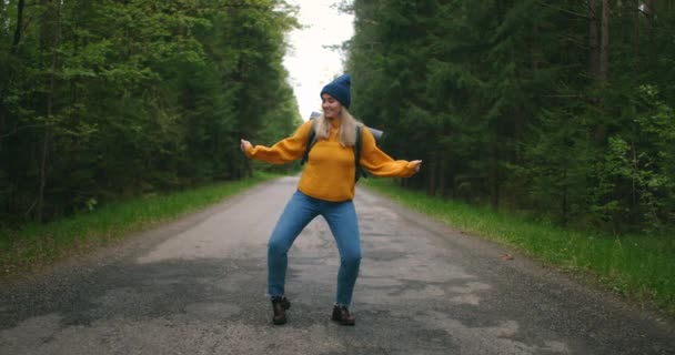Gelukkige vrouwelijke reiziger die grappig en leuk danst. Een vrouw in een gele trui en hoed met een rugzak op de weg in het bos. Een energieke vrouw met een rugzak. Het concept van open grenzen — Stockvideo
