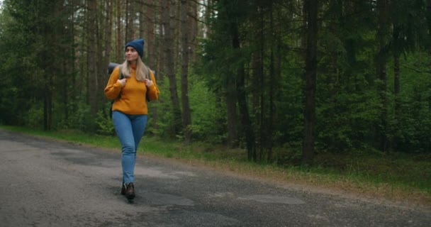 Vista frontal, planta principal: una mujer viajando a pie en un suéter amarillo y sombrero en una carretera forestal con una mochila y vistas a los árboles y la naturaleza. Solo en un viaje. El concepto de libertad y viajes. — Vídeos de Stock
