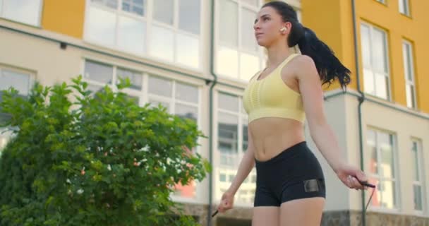 A young beautiful girl jumps on a skipping rope in a Park in slow motion in sports clothes. Play sports against the background of houses — Stock Video