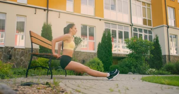 A young woman with headphones performs push-ups on a bench in a city park in slow motion. Training a young woman on a bench against the backdrop of the city and houses — Stock Video