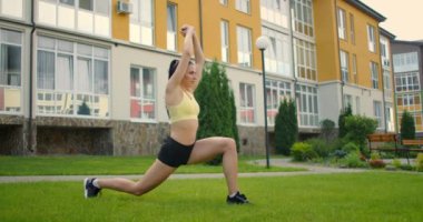 Parkta kulaklık takan genç bir kadın yoga yapıyor. Şehir parkındaki evlerin arka planında yavaş çekimde çimlerde yoga yapmak.