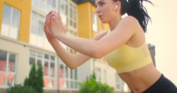 Uma jovem atleta com fones de ouvido realiza exercícios de salto em um parque da cidade contra o fundo dos edifícios. Salte em um banco no parque — Vídeo de Stock