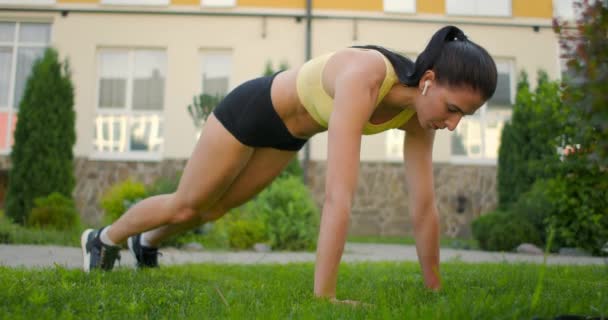 En kvinna med hörlurar tränar stående på gräset i en planka utför en bergsklättrare motion i en slow-motion system i en stadspark — Stockvideo