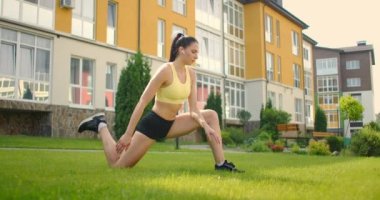 Kulaklıklı genç bir kadın, şehir parkında çimlerin üzerinde ağır çekimde esniyor. Spor yaparken müzik dinle.