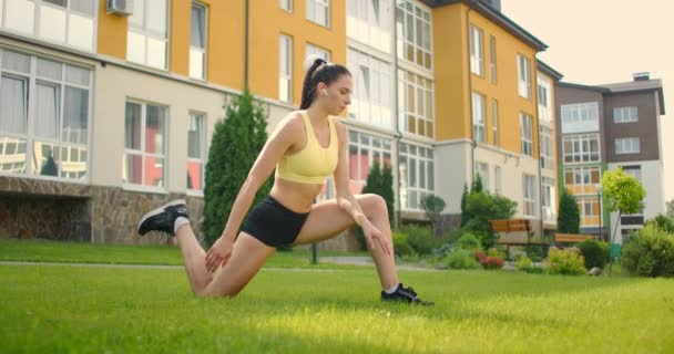 Una mujer joven con auriculares se está estirando en la hierba en un parque de la ciudad en cámara lenta. Escuchar música mientras practicas deportes — Vídeos de Stock