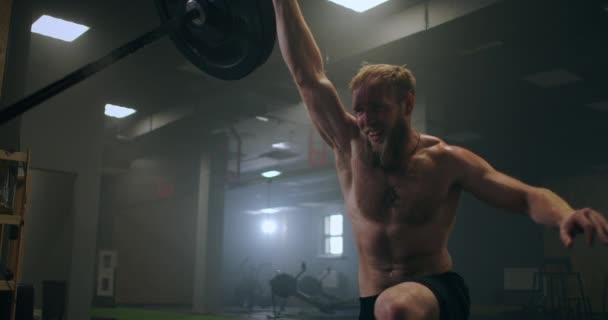 Pojken knuffar skivstången i gymmet med en hand. En atlet utan t-shirt i ett mörkt rum lyfter skivstången medan du tränar kraften i stöten — Stockvideo