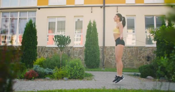 A young beautiful girl jumps on a skipping rope in a Park in slow motion in sports clothes. Play sports against the background of houses — Stock Video