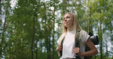 Ormanda ağaçların arasında sırt çantalı, düşünceli, ciddi bir kadın yürüyüşçünün el kamerasıyla çekilmiş fotoğrafı. Sırt çantalı genç bir kadın yaz günü ormanda yürür..