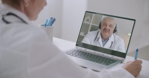 Videoconferência com médico chefe do hospital, mulher terapeuta está ouvindo o homem em exibição de seu laptop no escritório e fazendo anotações, escrevendo em papel — Vídeo de Stock