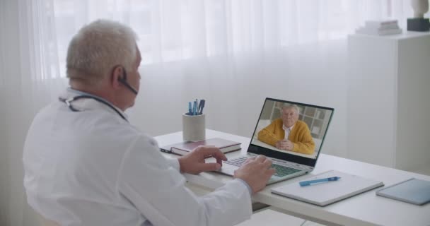 Консультации специалиста в области здравоохранения онлайн, старик звонит в больницу, чтобы поговорить с терапевтом, врач просматривает на ноутбуке — стоковое видео