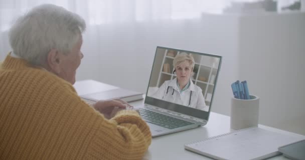 Old man is talking with his family doctor by web camera, consulting and advising, listening her, viewing at display of notebook — Stock Video