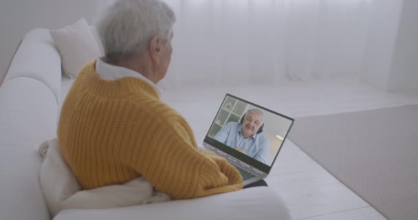 Anciano está sentado en el sofá en casa y charlando en línea con su hijo de mediana edad por videollamada, la comunicación a distancia de los familiares — Vídeos de Stock