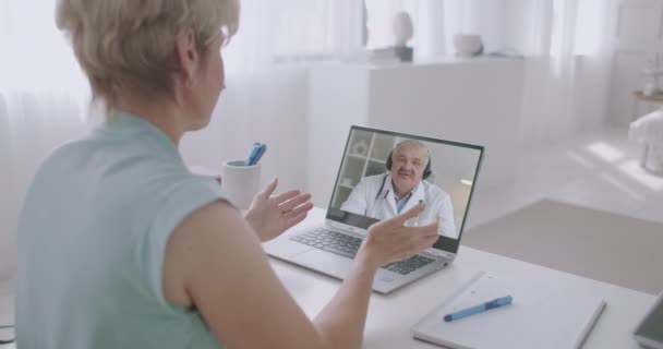 Wanita tua menonton webinar dokter terkenal melalui internet, menggunakan laptop di ruang tamunya, peduli tentang kesehatannya — Stok Video