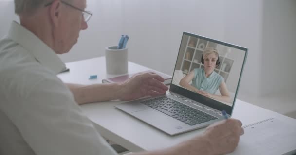 Anciano hombre está viendo en línea entrenamiento sentado en casa delante de la computadora portátil, mujer está dando conferencias, hombre está escribiendo notas — Vídeo de stock