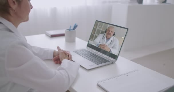 Consulta en línea de dos especialistas médicos por videollamadas en computadoras portátiles, la mujer está mirando a un colega masculino en la pantalla y escuchando — Vídeo de stock