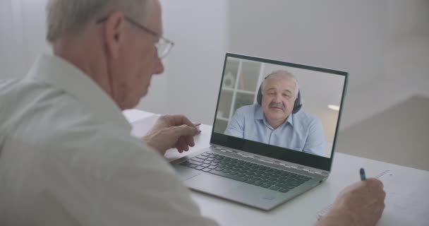 दोन पुरुष सहकारी व्हिडिओ कॉलद्वारे ऑनलाइन संप्रेषण करीत आहेत, बॉस बोलत आहेत आणि कर्मचारी लिहित आहेत — स्टॉक व्हिडिओ