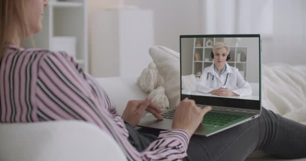 Молода жінка слухає онлайн-вебінар лікаря педіатра, сидячи вдома, використовуючи блокнот з інтернетом — стокове відео