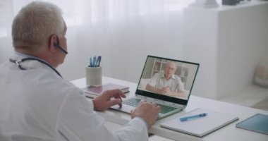 Online kardiyolog randevusu, yaşlı adam semptomlardan bahsediyor, erkek doktor hastanede otururken dinliyor.