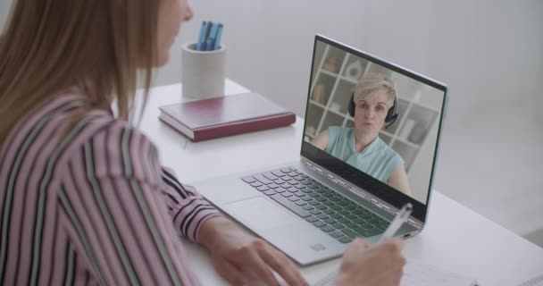 女性学生はノートパソコンでビデオチャットでプロジェクトの科学監督者と連絡を取り紙にメモを書き — ストック動画