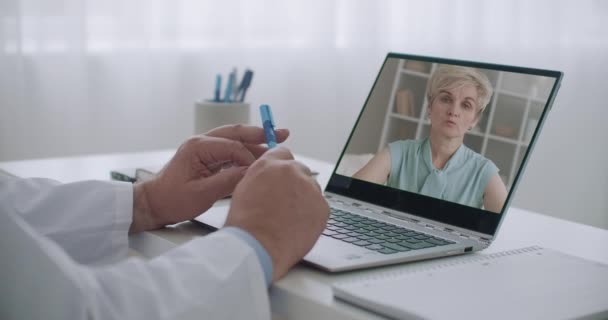 Mulher idosa está conversando com seu médico de família por videochamada, seu rosto na tela do laptop do médico no escritório da clínica — Vídeo de Stock