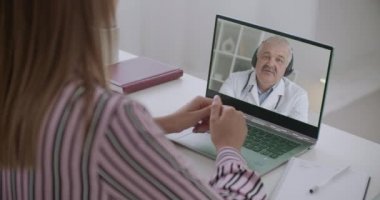 Orta yaşlı doktor dizüstü bilgisayarından kadın hastayla konuşuyor. Kadın evde oturan bir tıp uzmanı onu dinliyor.