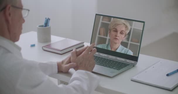 Bejaarde vrouw communiceert met dokter via webcamera, haar gezicht op laptop van arts, telegeneeskunde sessie — Stockvideo