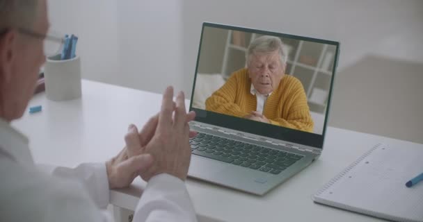 男性医師は患者の苦情を聞きオンラインチャットで連絡を取りノートパソコンのディスプレイで老人を見ています — ストック動画