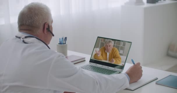 Het bijwonen van de arts luistert zijn oudere patiënt, communiceren via video bellen vanuit zijn kantoor in het ziekenhuis — Stockvideo