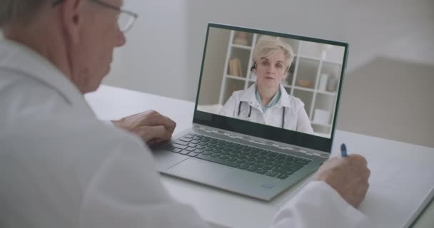 Online medicinsk vetenskaplig konferens, äldre läkare lyssnar kvinnliga professor genom bärbar dator och skriva anteckningar — Stockvideo