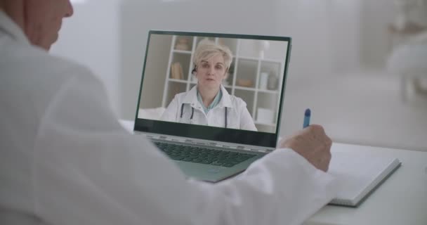 Онлайн-конференция медицинских работников, мужчина слушает женщину-физиолога и пишет заметки, лицо на экране ноутбука — стоковое видео