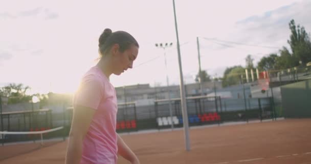 O jucătoare de tenis brunetă obosită merge de-a lungul terenului de tenis recuperându-se și concentrându-se. Pauză la un meci de tenis. Jucător de tenis după meciul de pe hartă la apusul soarelui în mișcare lentă . — Videoclip de stoc