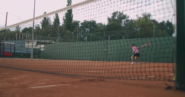 Slow motion komerční záběry tenisového tréninku přes tenisovou kurtní síť. Přímý pohled na ženskou atletku hrající tenis. Dospívající sportovec odpaluje míč během sportovního tréninku — Stock video
