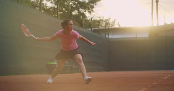 Uma jovem tenista morena joga uma bola ao pôr do sol em uma quadra de tênis. Uma mulher joga tênis profissional e dinamicamente em câmera lenta — Vídeo de Stock