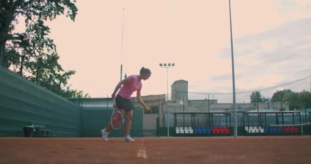 Movimento lento: esportista feminina durante sua prática. Um close-up de uma atleta feminina serve a bola de tênis. Jovem está batendo a bola com sua raquete de tênis ao pôr do sol — Vídeo de Stock