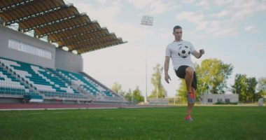 Futbol sahasındaki profesyonel erkek futbolcu spor ekipmanlarıyla ağır çekimde bir futbol topuyla jonglörlük yapıyor. Tribünlerin yanındaki stadyumda topu olan bir futbolcu.