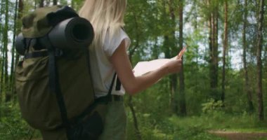 Elinde harita olan genç bir kadın ormanda ağır çekimde sırt çantasıyla dolaşıyor.