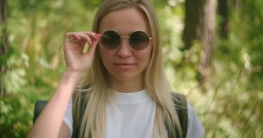 Güneş gözlüklü genç bir bayan gezgin kameraya bakar ve flört ederken gülümser. Sırt çantalı bir gezgin parkta ve ormanda ağır çekimde.
