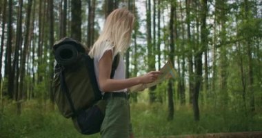 Elinde harita olan genç bir kadın ormanda ağır çekimde sırt çantasıyla dolaşıyor. Ormanda şortlu bir gezgin.