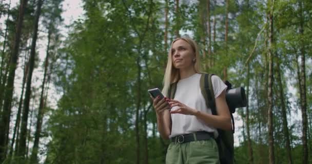 Una giovane donna con un cellulare cammina attraverso la foresta viaggiando con uno zaino al rallentatore. Viaggiatore in pantaloncini nel bosco in cerca di satelliti GPS — Video Stock