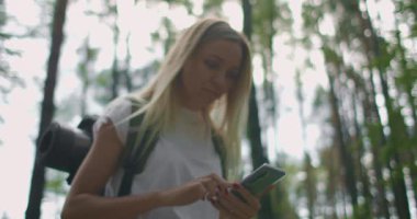 Cep telefonu olan genç bir kadın ormanda ağır çekimde sırt çantasıyla dolaşıyor. Cep telefonunuzdaki Navigator 'u kullanarak ormanda gezinin.